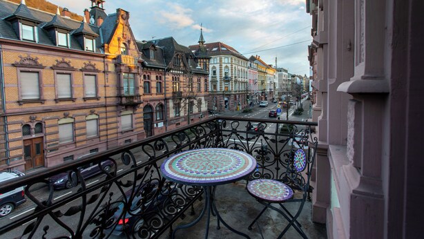 A balcony with wrought iron railings and two mosaic-topped tables, overlooking a street with historic buildings and a clear sky. - BoardingWorld Heidelberg (Heidelberg)