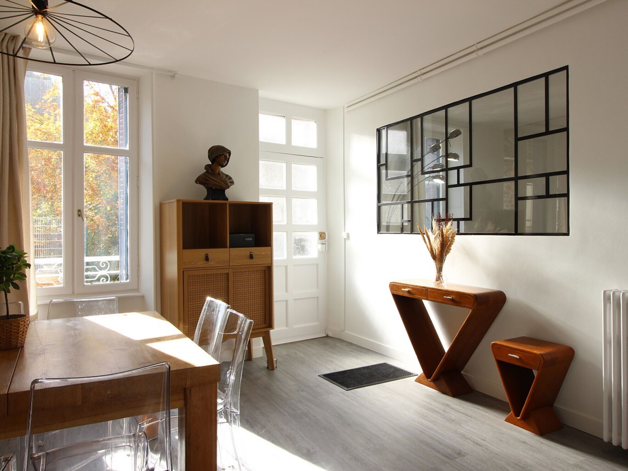 A modern dining area with a wooden table, clear chairs, a cabinet with a bust sculpture, and a wall-mounted mirror.