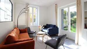 Ett modernt vardagsrum med en soffa, en fåtölj och ett soffbord. Det finns stora fönster med gardiner, ett element och en bokhylla.