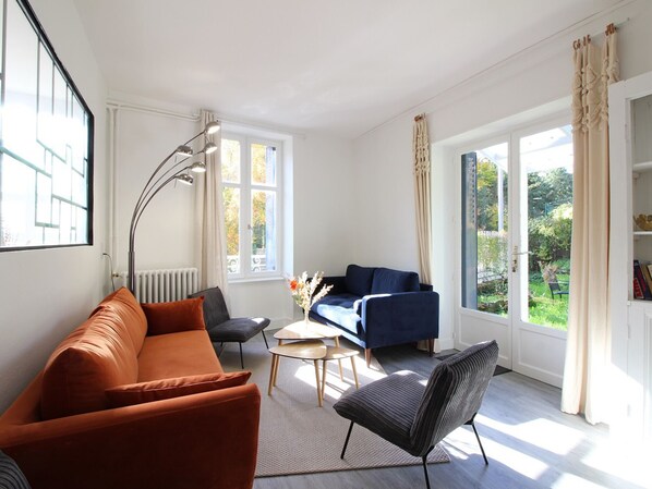 Un salon moderne avec un canapé, un fauteuil et une table basse. De grandes fenêtres sont présentes, ainsi que des rideaux, un radiateur et une bibliothèque.