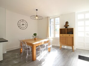 Un coin repas avec une table en bois, des chaises en plastique transparent, une horloge murale, un luminaire suspendu et un buffet en bois sur lequel repose un buste.
