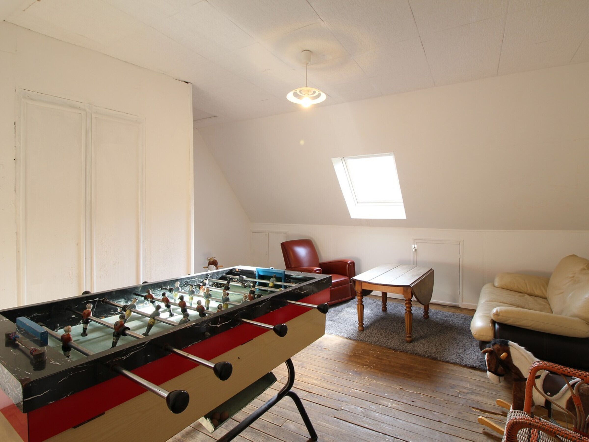 Una habitación con mesa de futbolín, una silla roja, una mesita, un sofá y una ventana.