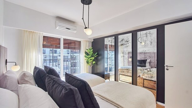 Un dormitorio moderno con una cama grande, una mesita de noche, una lámpara y vistas de la ciudad a través de un ventanal corredero.