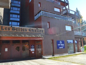 Um edifício de vários andares com fachada de madeira e varandas. Há placas com os dizeres 'Les Belles Challes' e 'Les Arcs Agency'.
