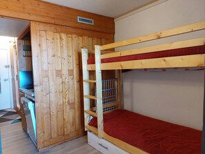 Uma cama de solteiro com colchão vermelho, paredes de madeira, uma pequena cozinha e uma porta que dá para outro quarto.