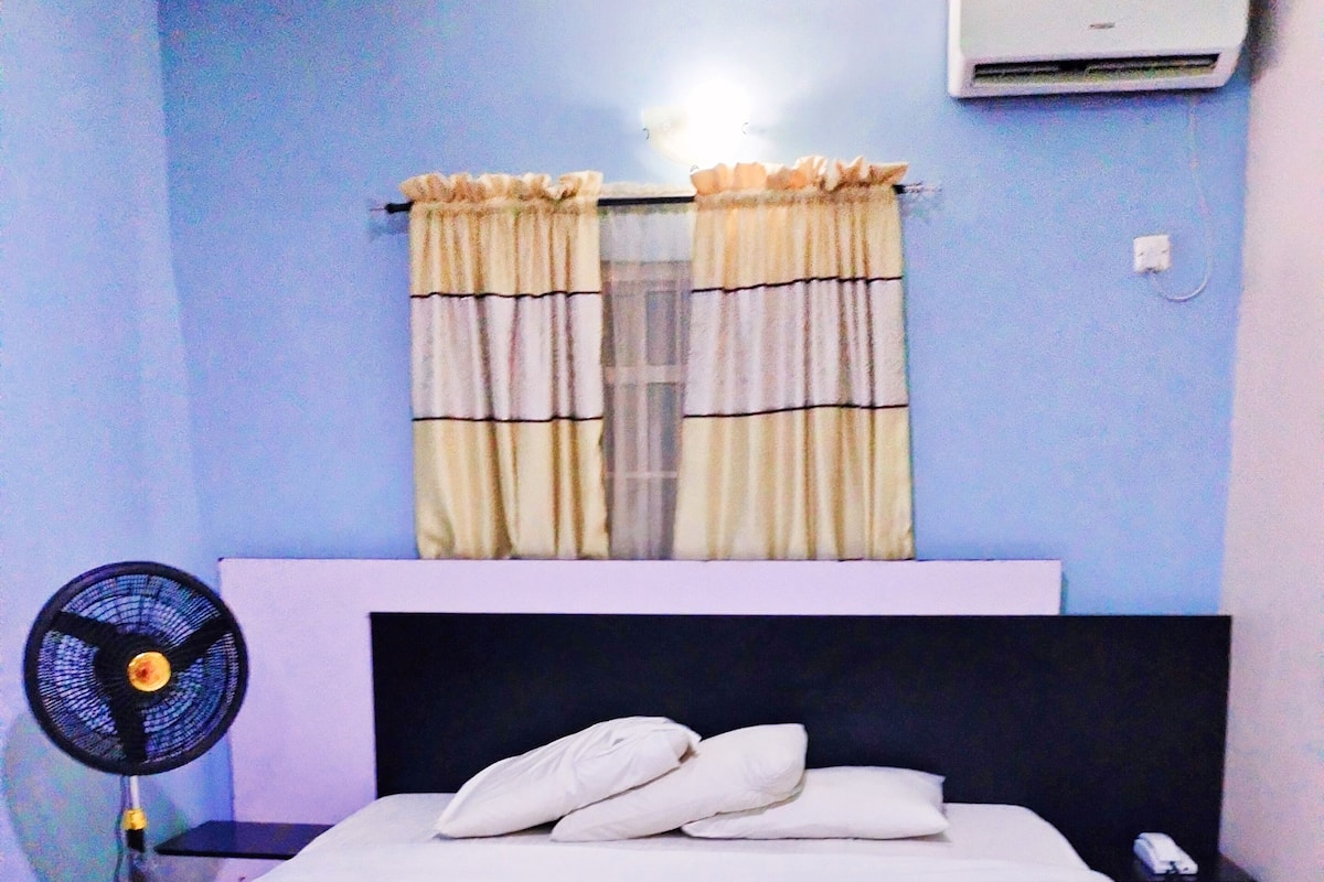 Quarto com cama, mesa de cabeceira, ar-condicionado e ventilador.