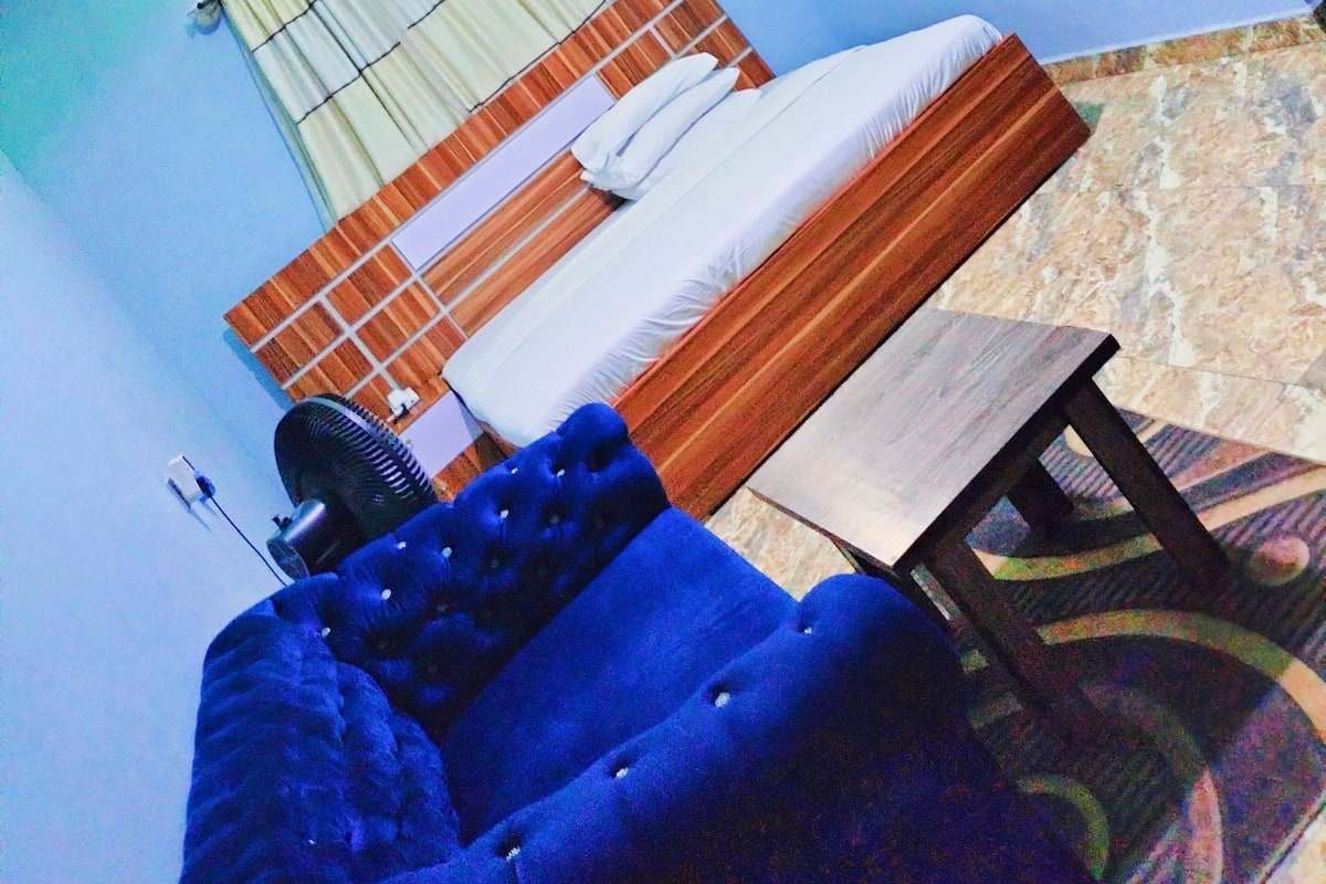 Quarto com cama, poltrona azul estofada, ventoinha e ar condicionado fixado na parede.