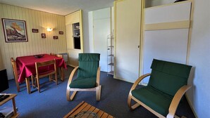 A room with a dining table and chairs, a green cushioned chair, a wooden coffee table, and a door leading to another room. - Ski-in/Ski-out Apartment in Reberty 1850, Les Menuires - Sleeps 4, 1 Bed, 1 Bath (Les Menuires)