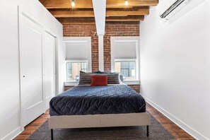 A bedroom with a bed, a bench, a window, and a brick wall. - Oak St. Loft #6- 1bed + Walk to Downtown + Parking (Kansas City)