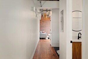 A narrow hallway with wooden flooring, white walls, and a brick wall in the background. - Oak St. Loft #6- 1bed + Walk to Downtown + Parking (Kansas City)