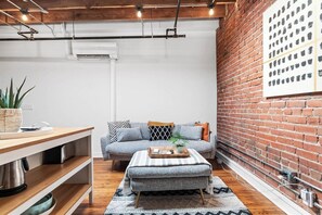 A cozy living area with a sofa, ottoman, and a dining table with a plant. - Oak St. Loft #6- 1bed + Walk to Downtown + Parking (Kansas City)