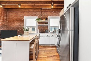 Une cuisine avec un mur en briques, un plafond en bois, des armoires blanches, un réfrigérateur en acier inoxydable et un îlot central en bois doté de sièges.