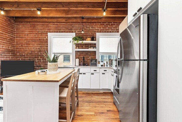 A kitchen with a brick wall, wooden ceiling, white cabinets, a stainless steel refrigerator, and a wooden island with seating.