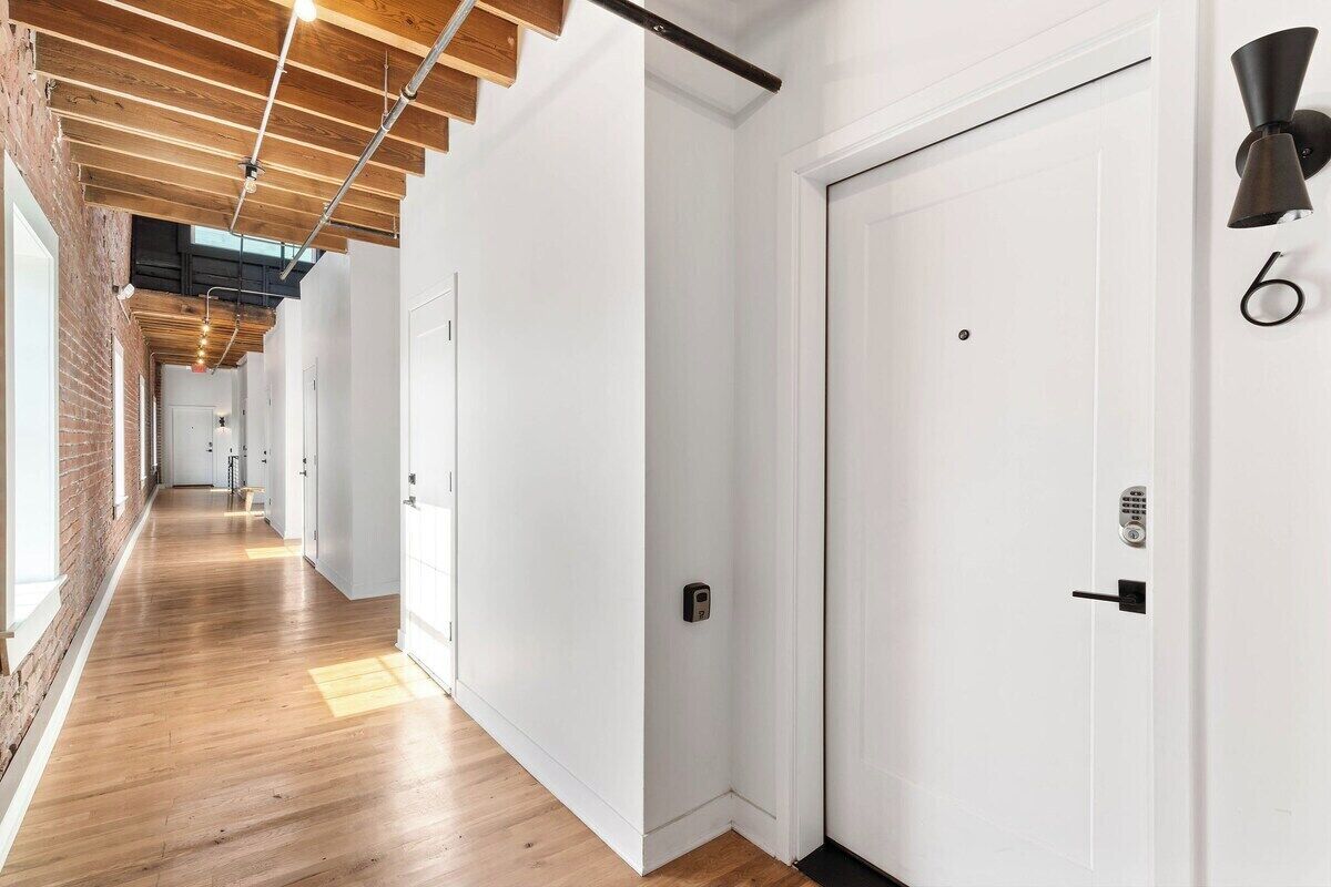 A hallway with wooden floors, white walls, and a ceiling with exposed beams. There are doors with numbers on them, and a wall-mounted light fixture.