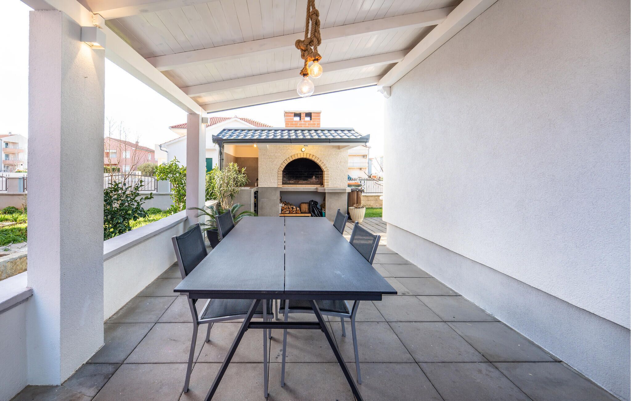 An outdoor patio with a long table and chairs, a brick oven, and a hanging light fixture.