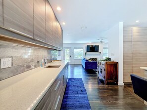 Une cuisine moderne dotée d’un four encastré, d’un grand îlot central avec un évier, et d’un tapis bleu qui mène à un espace de vie comprenant un canapé et un téléviseur.