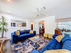 A modern living room with a blue sofa set, a glass coffee table, and a ceiling fan. - Modern Coastal Home Near Las Olas + Heated Pool (Fort Lauderdale)
