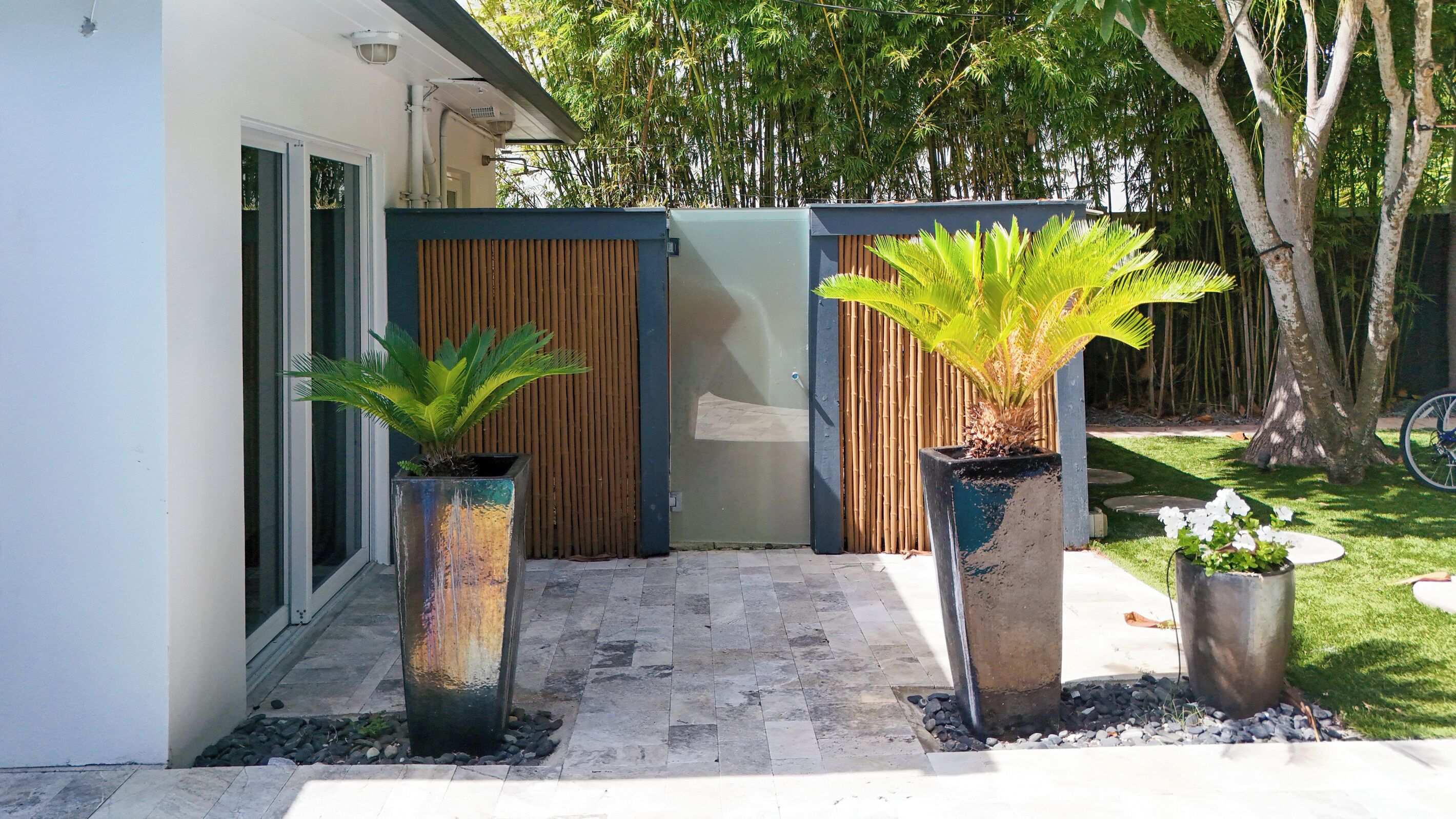 A modern outdoor entrance with a concrete pathway leading to a wooden gate, flanked by two large planters with tropical plants.