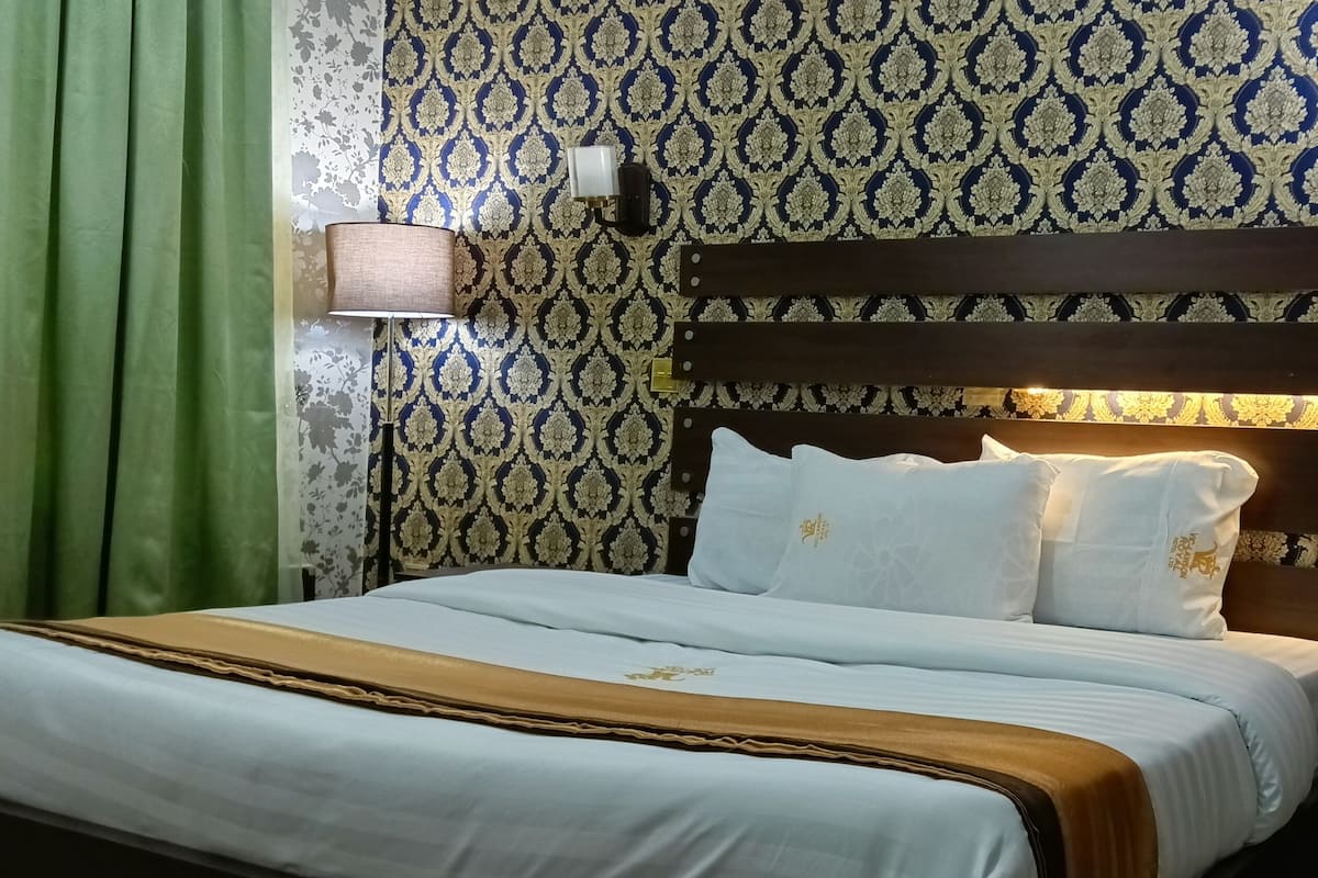 Hotelkamer met een bed, groene gordijnen, behang met een patroon, een nachtkastje met telefoon en wandlampen.