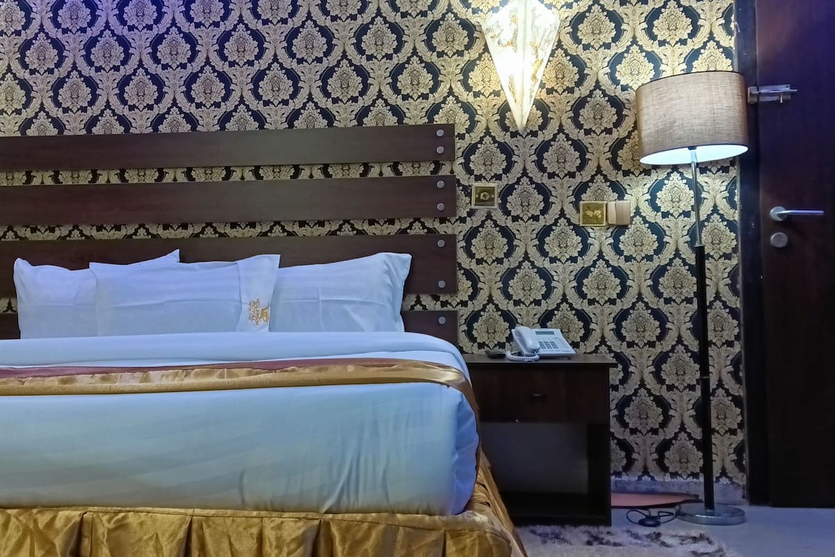 Een keurig opgemaakt bed met een houten hoofdbord, een nachtkastje met een lamp en behang met een patroon.