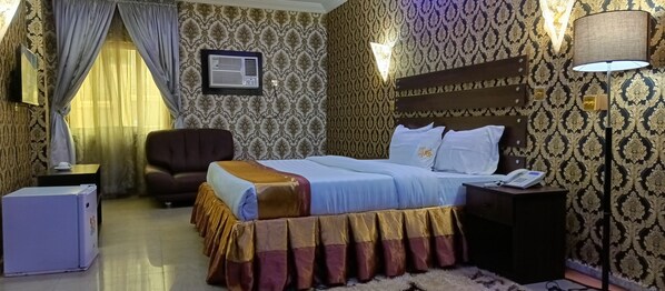 A hotel room with a bed, a desk, a chair, a lamp, and a wall-mounted air conditioner.