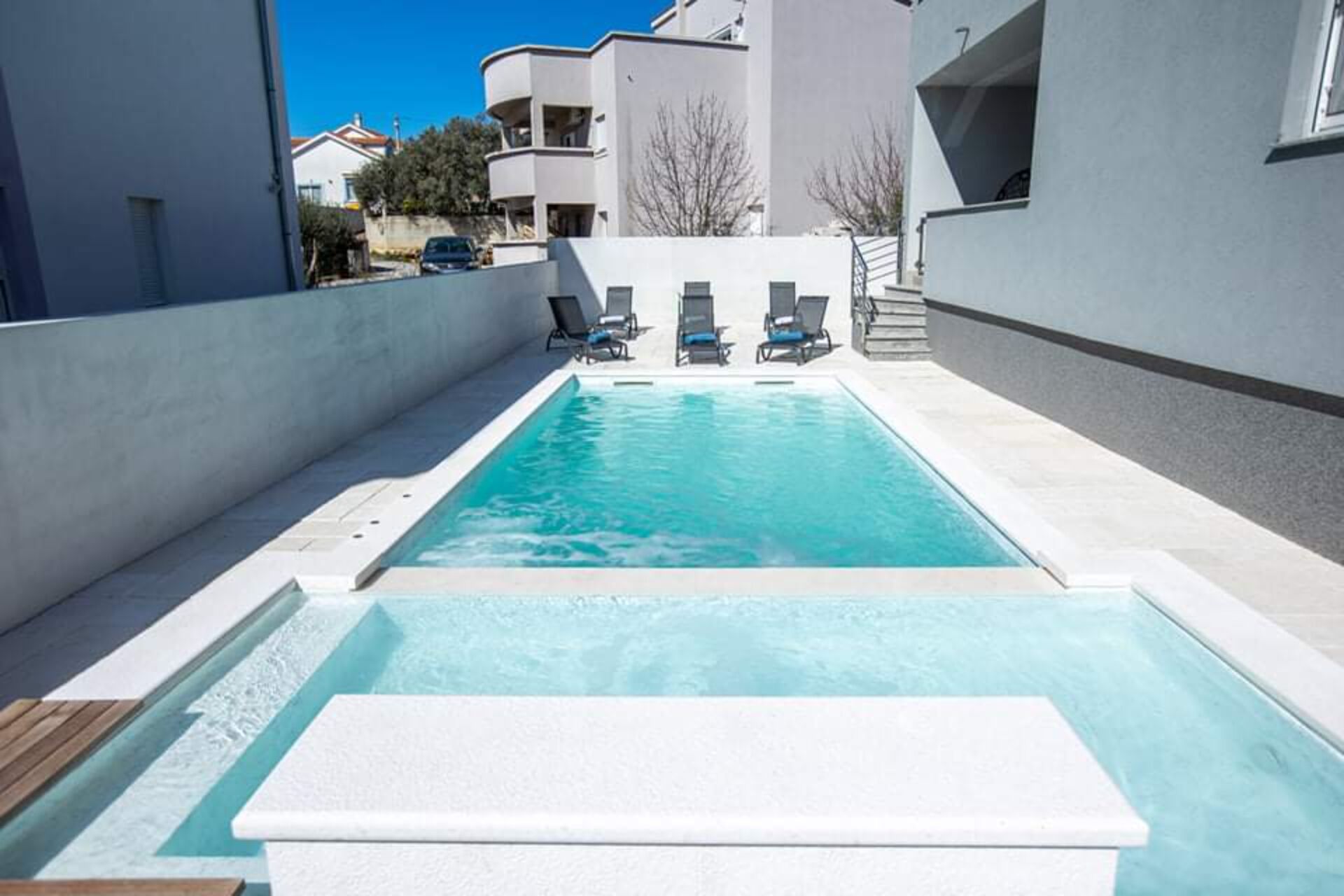 A rectangular swimming pool with a white deck in the foreground, surrounded by a low wall. There are three lounge chairs aligned along the wall, and a staircase leading to an upper level.