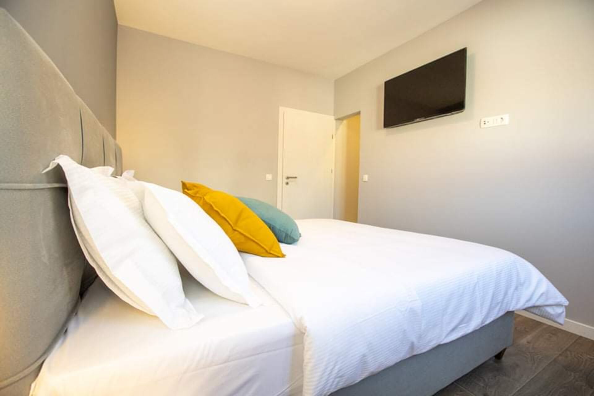 A bedroom with a bed, pillows, a television on the wall, and a door in the background.