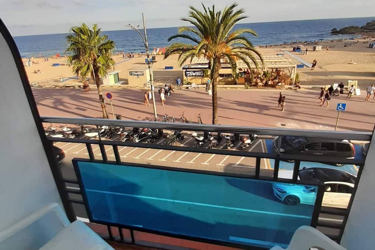 Een balkon met twee witte plastic stoelen, een blauwe pooltafel en uitzicht op een strand met palmbomen en een heldere lucht.