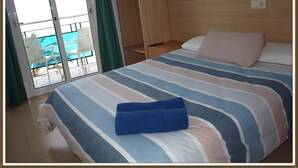 A hotel room with a bed, a wooden wardrobe, a balcony with a chair, and a pool view.