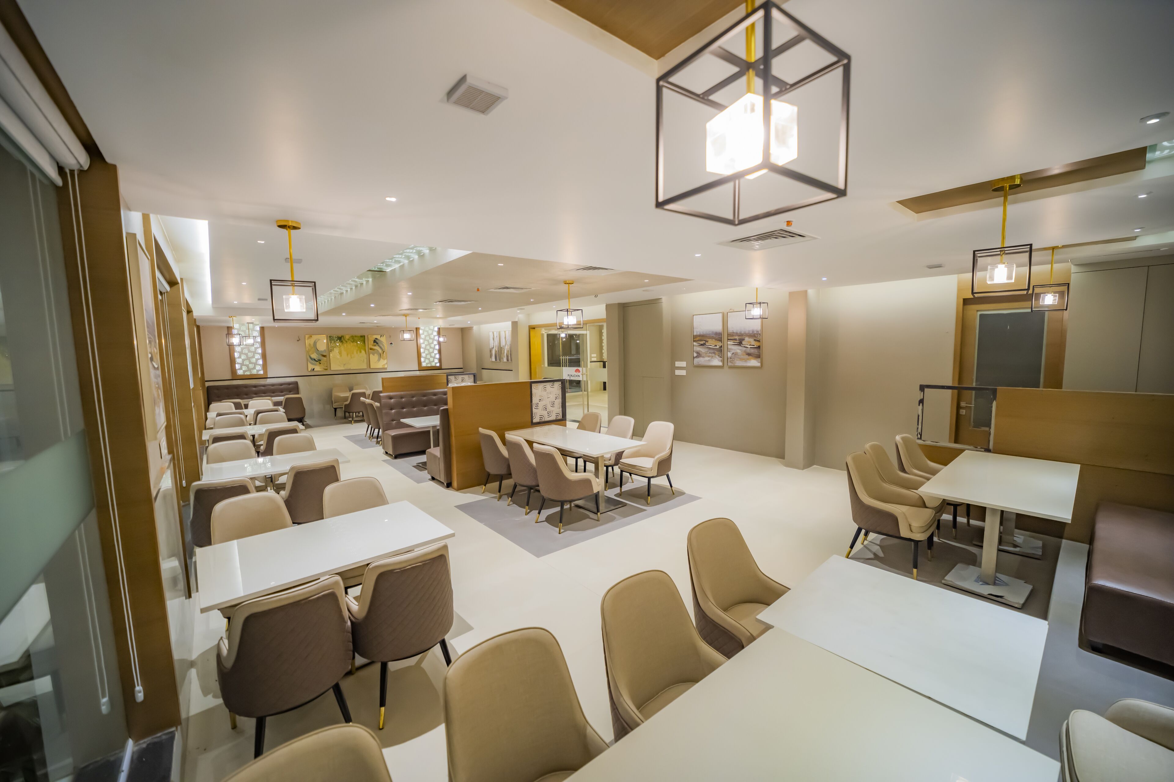 A modern hotel dining area with multiple tables and chairs, a central seating area, and contemporary lighting fixtures.