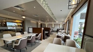 Un comedor moderno de hotel con mesas y sillas largas, una cocina al fondo y ventanas grandes con vistas.
