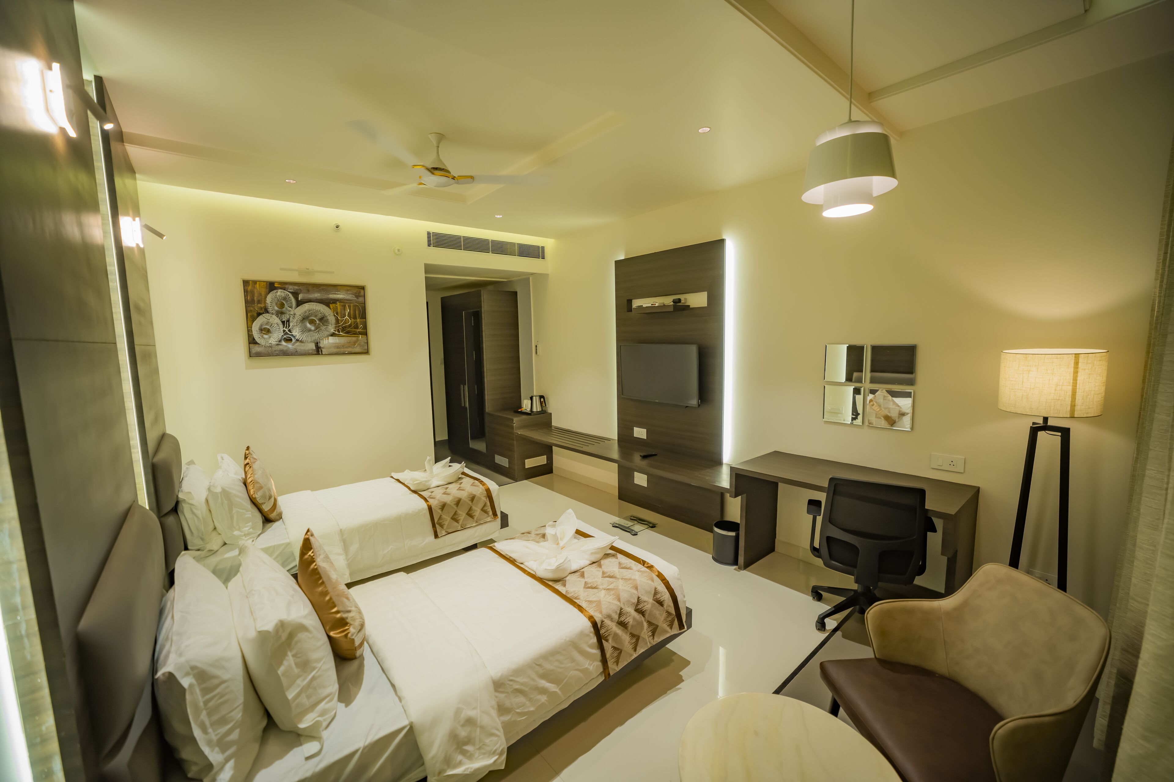 A hotel room with two beds, a desk, a chair, a TV, and a ceiling fan.