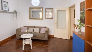 Una sala de estar con un sofá, una mesita redonda, un lámpara colgante, un espejo y una puerta que da a otra habitación.