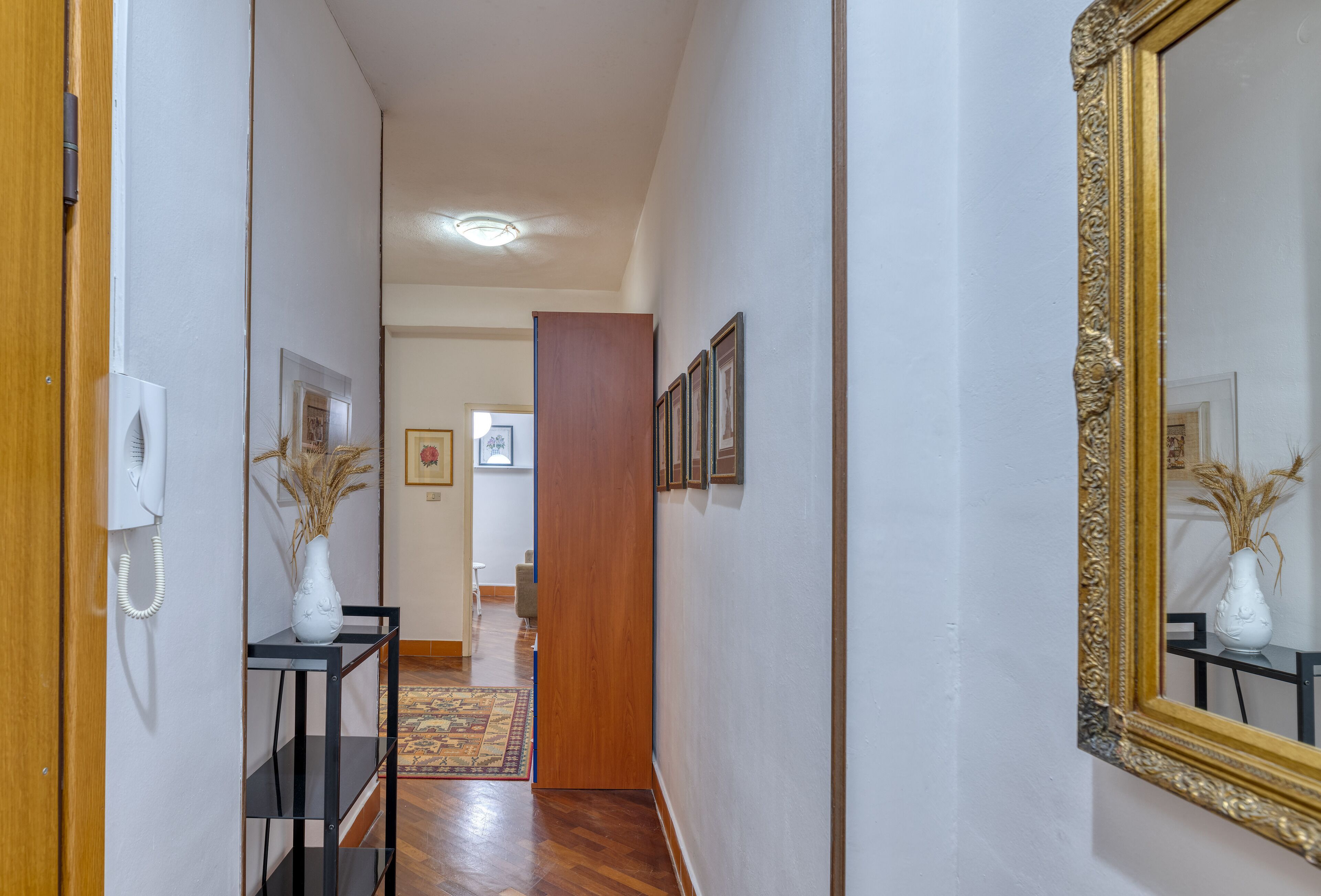 A hallway with a wooden door, a white vase, framed pictures, and a mirror with a gold frame.