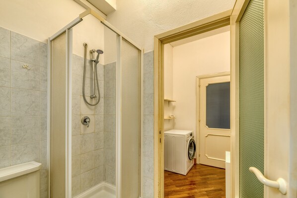 A bathroom with a shower, toilet, and a washing machine in the adjacent room. - IzzHome Sonnino (Cagliari)