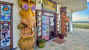 Un bâtiment dont la façade est ornée d’une grande fresque colorée représentant un visage, une devanture de magasin avec une enseigne indiquant « Airbrush », et une vue sur l’océan.