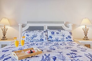 A bedroom with a bed, two bedside lamps, a tray with fruit and drinks, and a pillow with a beach theme.