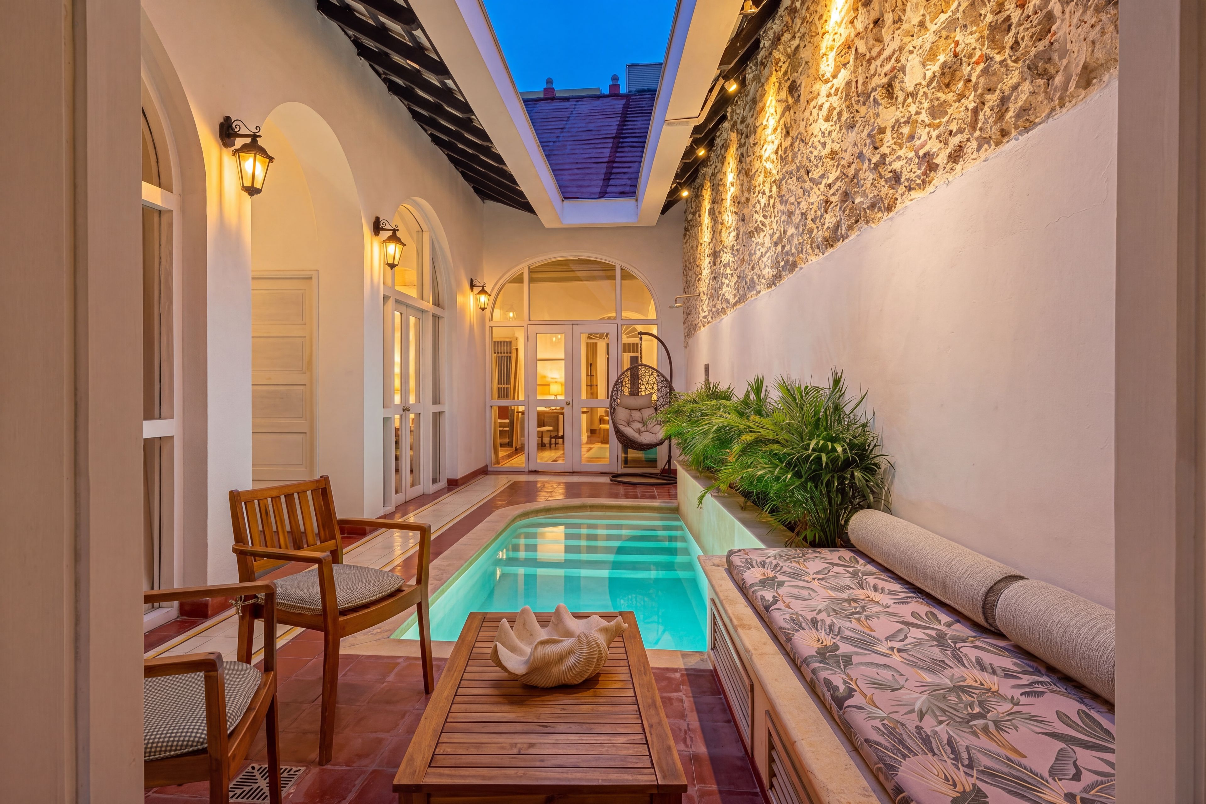 A pool area with a wooden table and chairs, a cushioned bench, and a stone wall with lights.