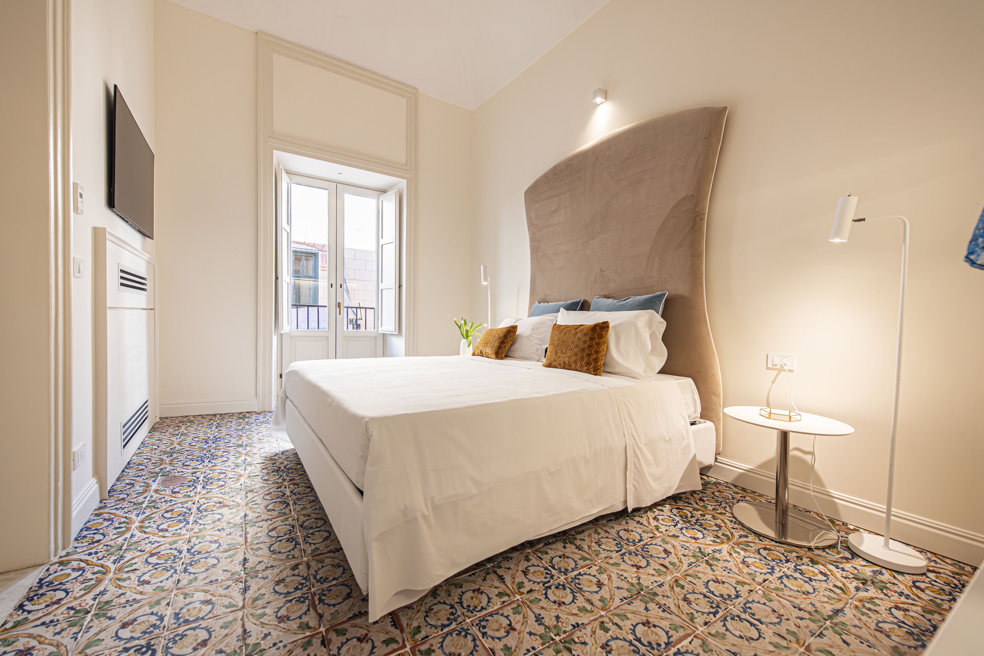 A hotel room with a bed, a TV mounted on the wall, a small table with a lamp, and patterned floor tiles.
