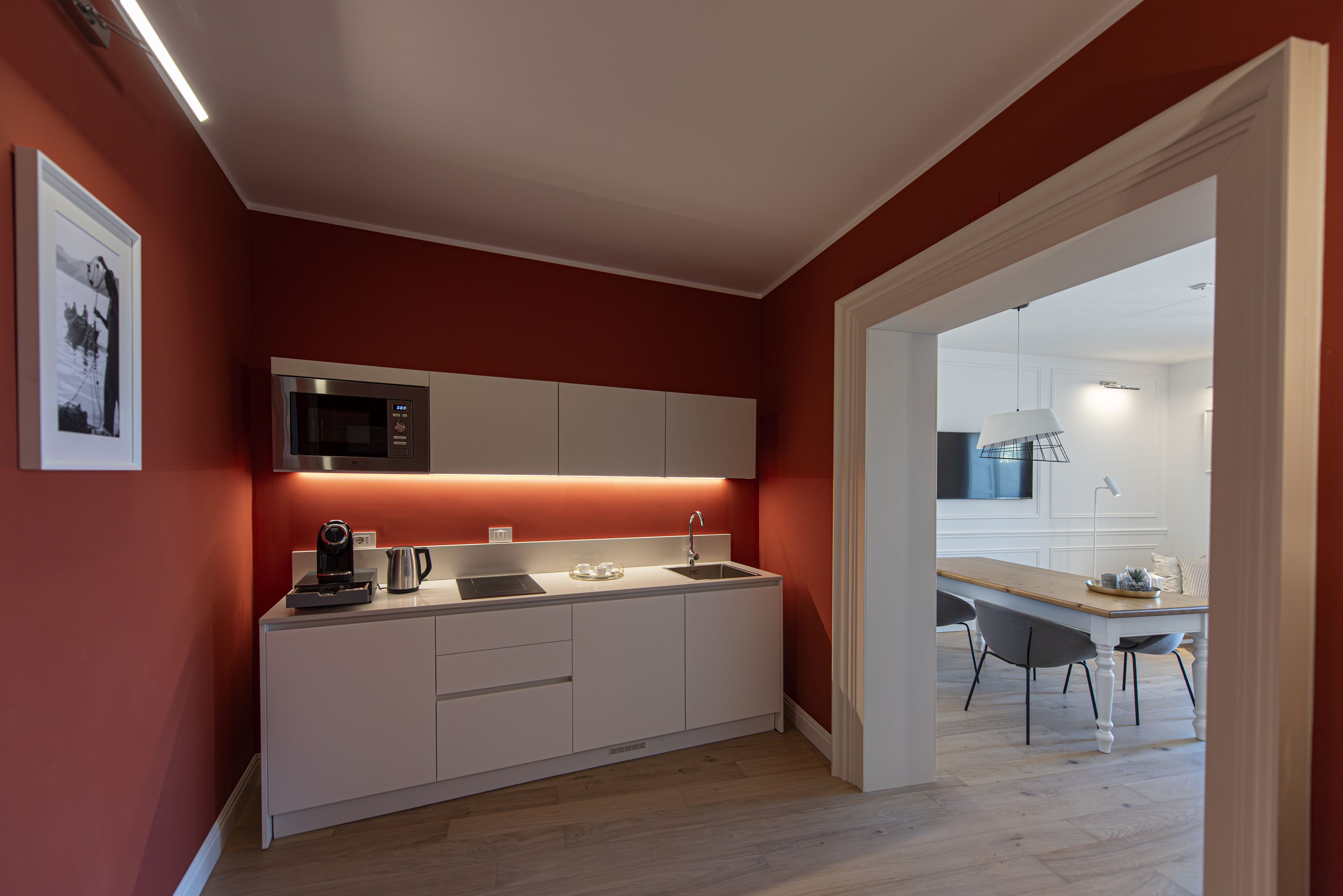 Una cocina moderna con armarios blancos, microondas y cafetera. Se ve un comedor con mesa y sillas a través de un arco.