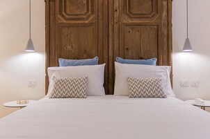 A neatly made bed with white linens and patterned pillows, a wooden wardrobe behind it, and a small bedside table with a lamp.