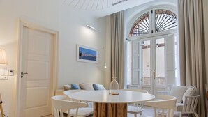 Una habitación con una mesa redonda de madera rodeada de sillas blancas, una puerta blanca, un cuadro enmarcado en la pared y un amplio ventanal con cortinas.