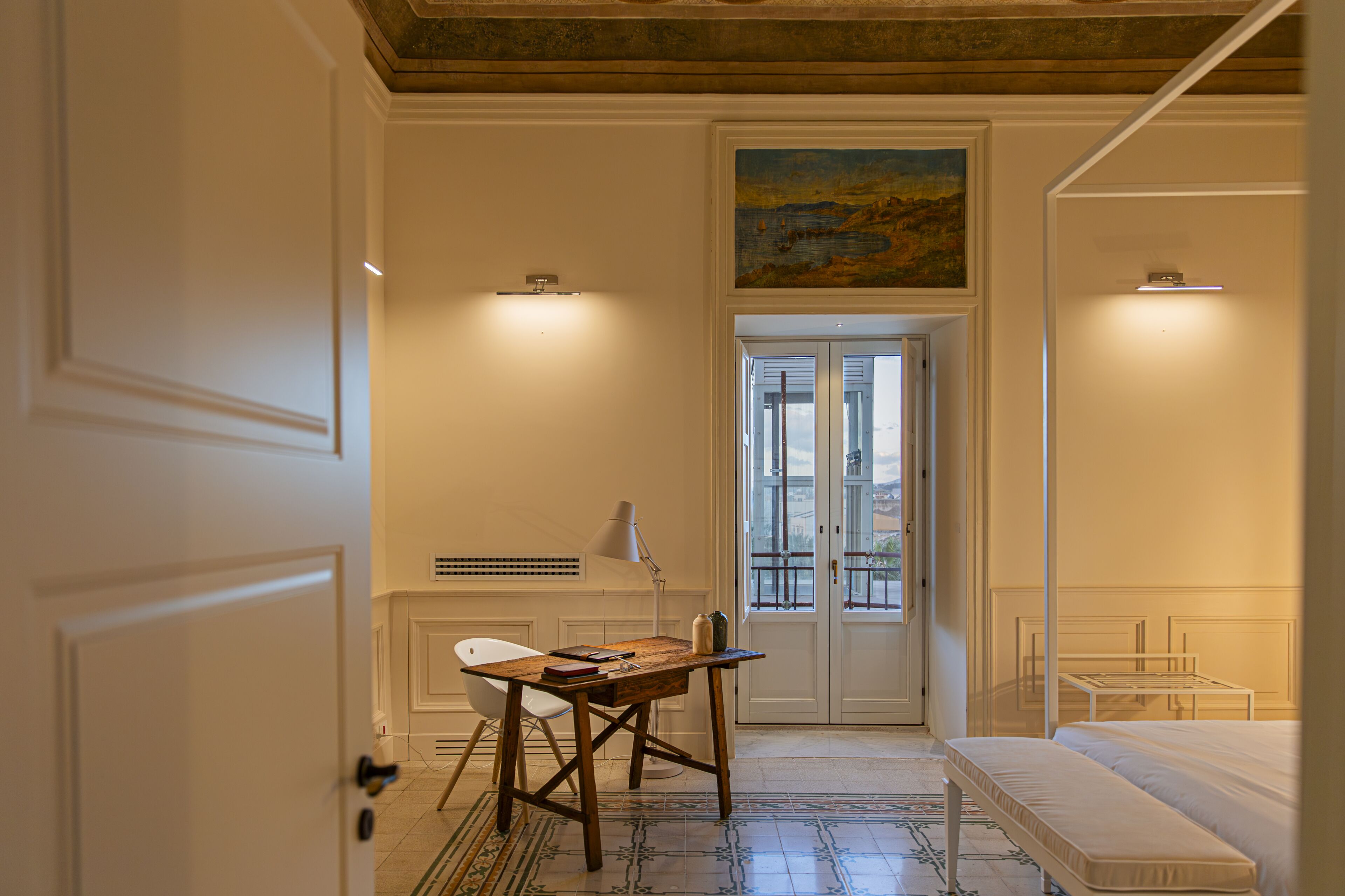 A room with a wooden desk, a white chair, a bed, and a painting on the wall.