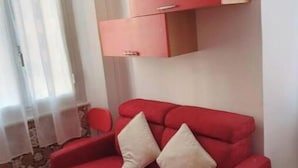 Un canapé rouge avec deux coussins, un meuble en bois et une chaise rouge dans une pièce dont les murs sont recouverts d’un papier peint à motifs et dont le sol est en bois.