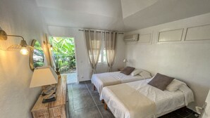 A bedroom with two beds, a wooden nightstand, a lamp, and a view of greenery outside.