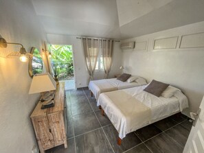Quarto com duas camas, uma mesinha de cabeceira de madeira, uma luminária e vista para o verde lá fora.