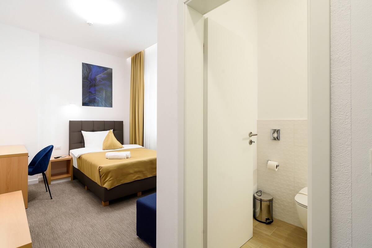 A hotel room with a bed, a desk, a chair, a small table, a trash bin, and a bathroom with a toilet and sink.