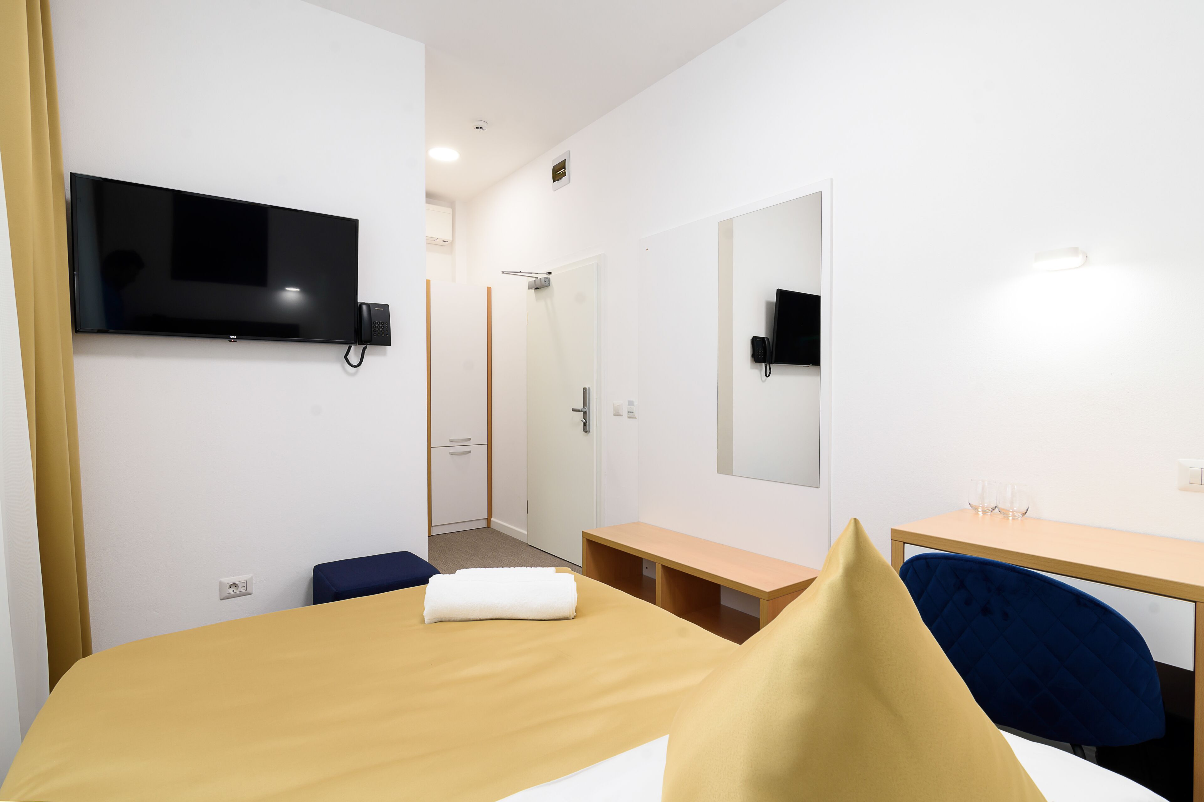 A hotel room with a bed, a desk, a chair, a TV mounted on the wall, and a mirror.