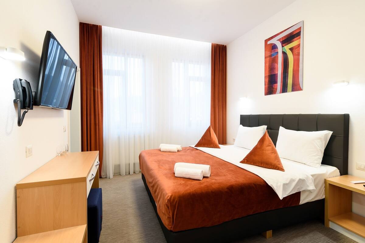 A hotel room with a bed, a desk, a chair, a TV, and a painting on the wall.