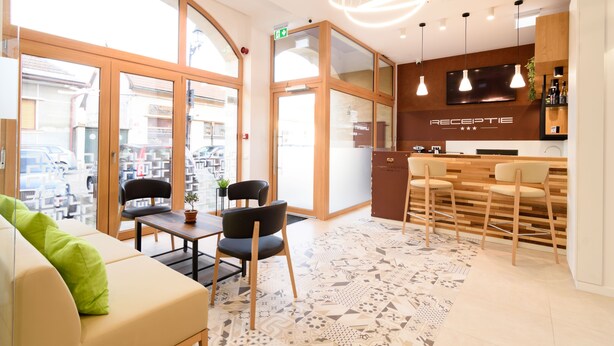 Un área de recepción moderna de hotel con un mostrador de madera, dos taburetes de bar, un sofá con un cojín verde y una alfombra estampada.